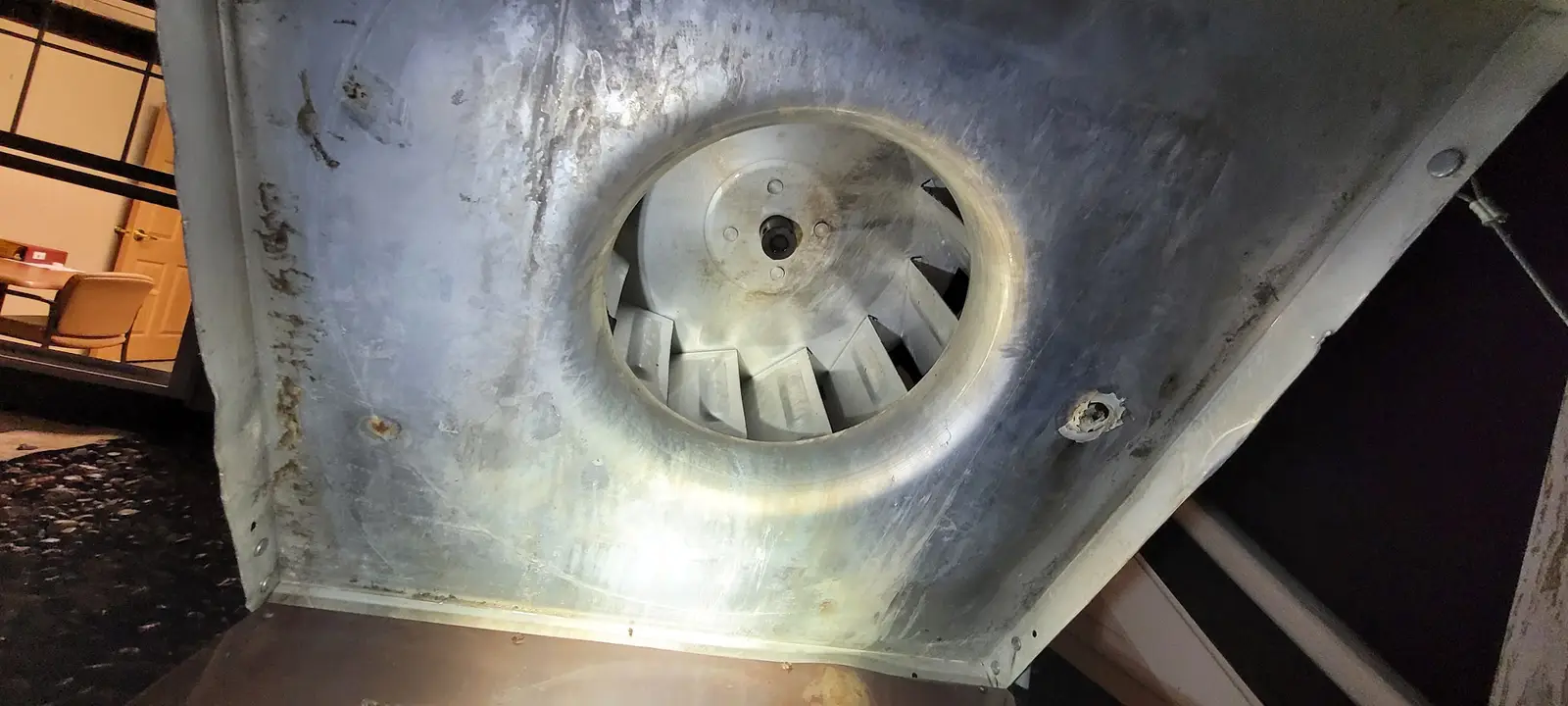Exhaust fan scroll seen from inside a kitchen hood plenum after cleaning, a lit room through the window behind it